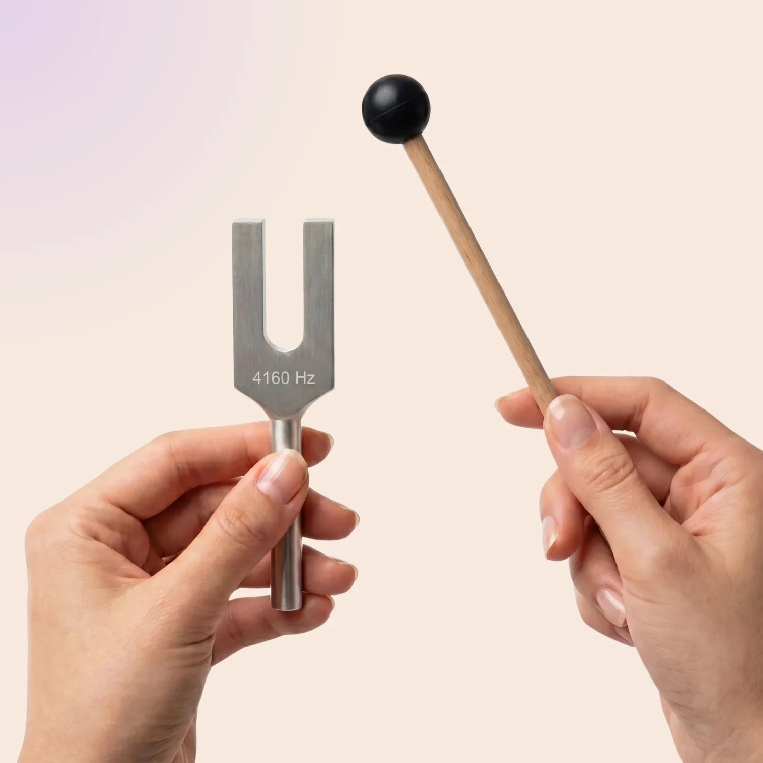 A metal tuning fork labeled 4160 Hz held in a hand, paired with a wooden mallet featuring a black rubber tip, against a soft beige gradient background.