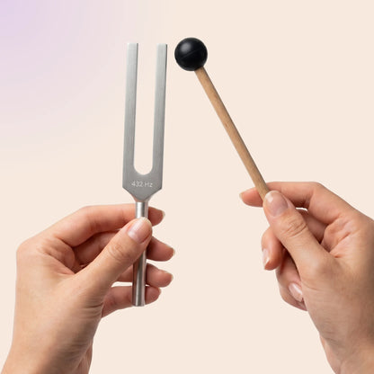 432 Hz tuning fork with a mallet held by hands against a light pink background