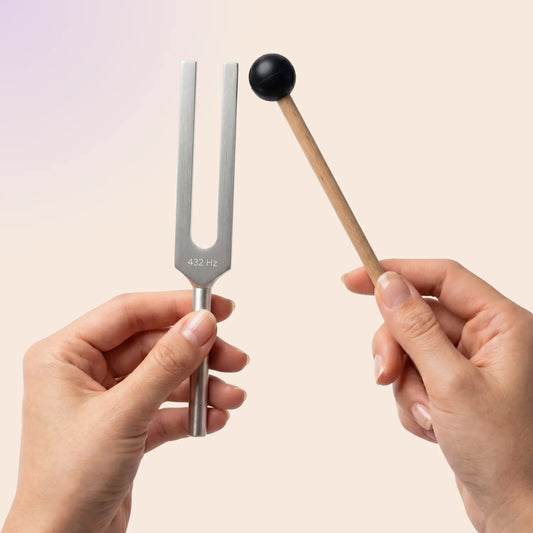 432 Hz tuning fork with a mallet held by hands against a light pink background