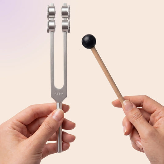 A stainless steel tuning fork labeled '64 Hz' held by a hand, alongside a wooden mallet with a black rubber tip held by another hand, against a soft beige background.