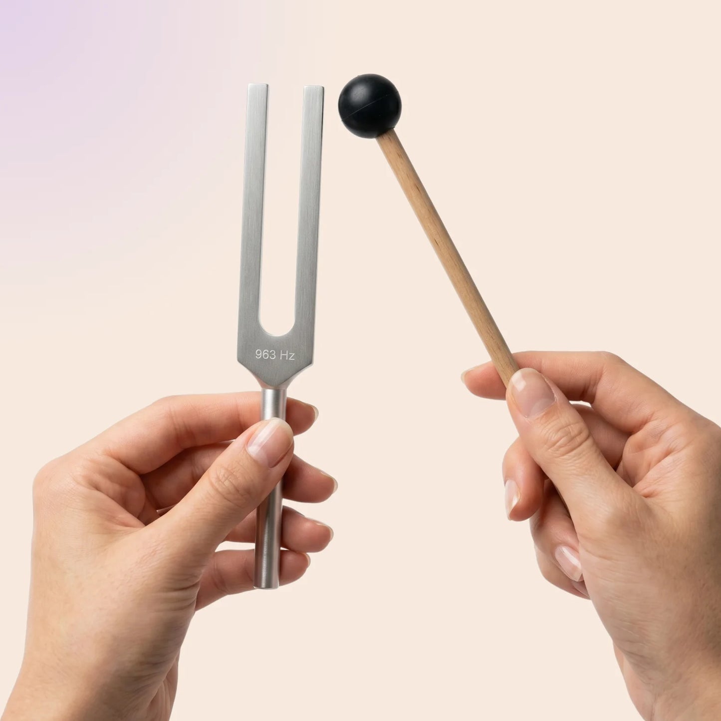 963 Hz tuning fork held by a hand with a mallet, on a light pink background